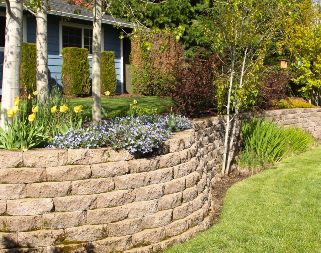 Curved block retaining wall with colorful flowers, shrubs, and a green lawn - Cajun Care Demolition & Tree Service in New Orleans
